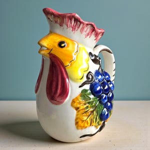 Op de afbeelding: Een keramische kan in de vorm van een haan, met een gele kop, rode kinlellen en een witte body. Versierd met blauwe druiven en gele bladeren. De rand is beschilderd met rode en witte accenten.