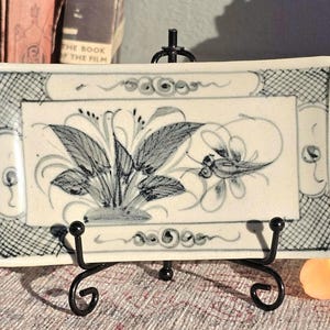 May include: Rectangular ceramic dish with a hand-painted floral design in blue and white. The dish is displayed on a black metal stand. The floral design features stylized leaves and a flower. The border has a crosshatch pattern.