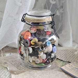May include: A clear glass jar filled with a variety of colorful buttons. The jar has a metal clasp and a rope-wrapped lid. The buttons are in various sizes, shapes, and colors, including red, blue, yellow, and white. The jar sits on a surface with vintage papers.