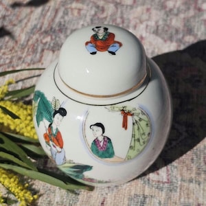 Puede incluir: Un tarro de jengibre de cerámica blanca con tapa, con figuras y escenas asiáticas pintadas a mano en colores vibrantes. El tarro tiene un borde dorado y una pequeña figura en la tapa. Una pieza decorativa para el hogar.
