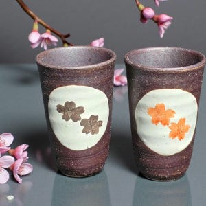 May include: Two brown ceramic cups with a textured finish. Each cup has a white oval with two cherry blossom designs, one in brown and the other in orange. Pink cherry blossoms are in the background.