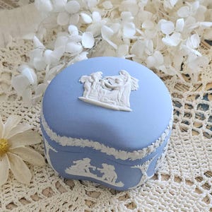 May include: A light blue, heart-shaped trinket box with white bas-relief figures and floral trim. The lid features a central relief scene. The box is set against a backdrop of white flowers and lace.