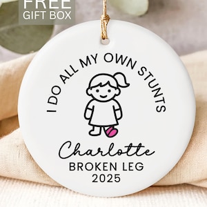 May include: White ceramic ornament with the text "I DO ALL MY OWN STUNTS" around a cartoon girl with a broken leg. The name "Charlotte" and "BROKEN LEG 2025" are below the illustration. Includes a gold string for hanging.