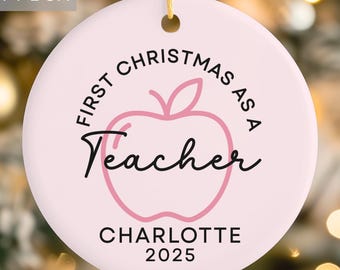 May include: A round, light pink Christmas ornament with the text "First Christmas as a Teacher" in black script. A pink apple graphic is in the center. The name "Charlotte" and the year "2025" are below the apple. A "Free Gift Box" label is in the upper left.