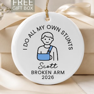 May include: A round white ceramic ornament featuring a black line drawing of a person in a blue arm sling. The text reads: I DO ALL MY OWN STUNTS, Scott, BROKEN ARM, 2026. The ornament hangs from a gold string against a neutral background.