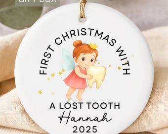 May include: White ceramic Christmas ornament with a gold string. The ornament features a cartoon fairy holding a tooth, with the text "First Christmas With A Lost Tooth Hannah 2025". Includes a "FREE GIFT BOX" label.