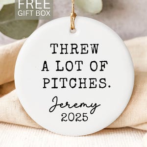 May include: A white ceramic ornament with the text "THREW A LOT OF PITCHES. Jeremy 2025" in black. The ornament has a gold string for hanging and is labeled "FREE GIFT BOX".
