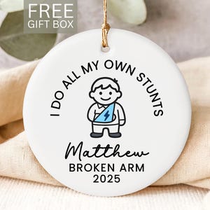 May include: White ceramic ornament with a gold string for hanging. The ornament features a cartoon illustration of a person with a broken arm, the text "I DO ALL MY OWN STUNTS", the name "Matthew", and "BROKEN ARM 2025".