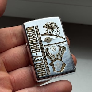 May include: A silver Harley-Davidson lighter with gold-colored accents. The lighter features an eagle graphic and the text "HARLEY-DAVIDSON BORN TO RIDE FORCED TO WALK". The design includes an engine illustration.