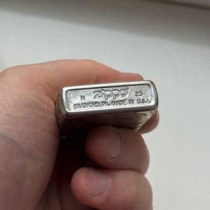 May include: A silver Zippo lighter with the brand name and "Made in USA" text. The lighter has a rectangular shape and is held in a hand. The text "25" is also visible.