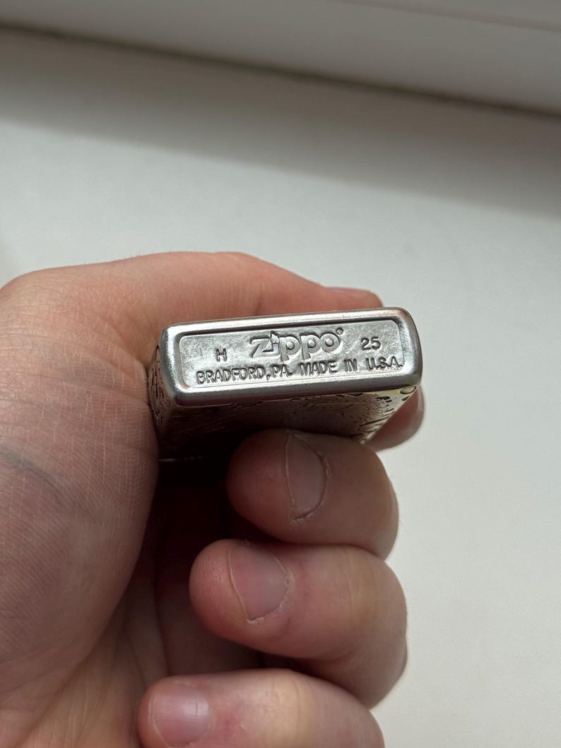 May include: A silver Zippo lighter with the brand name and "Made in USA" text. The lighter has a rectangular shape and is held in a hand. The text "25" is also visible.
