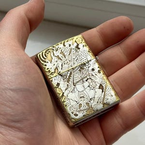 May include: A gold and silver-toned lighter with an intricate engraved design featuring a warrior and dragon motif. The lighter is rectangular and held in a hand, showcasing the detailed artwork.