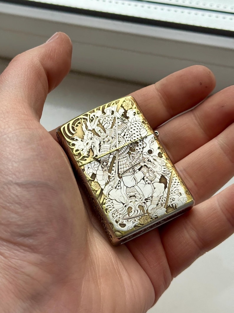 May include: A gold and silver-toned lighter with an intricate engraved design featuring a warrior and dragon motif. The lighter is rectangular and held in a hand, showcasing the detailed artwork.