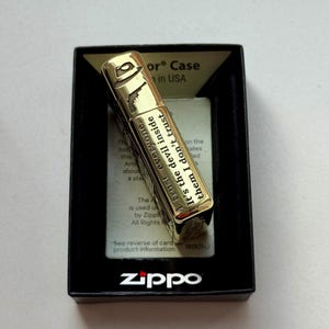 Pu&ograve; includere: Accendino Zippo color oro con un design a fedora e pistola, presentato in una scatola nera. L'accendino presenta il testo "I trust everyone. It's the devil inside them I don't trust."