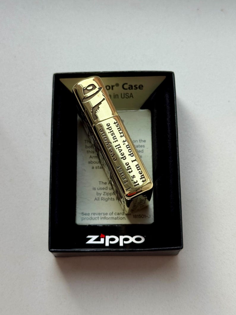 Pu&ograve; includere: Accendino Zippo color oro con un design a fedora e pistola, presentato in una scatola nera. L'accendino presenta il testo "I trust everyone. It's the devil inside them I don't trust."