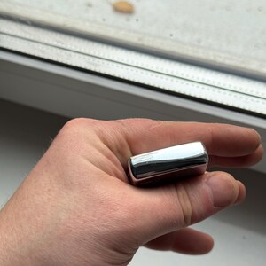 May include: A shiny, rectangular, chrome-colored lighter held in a hand. The lighter has rounded edges and reflects the surrounding environment. The lighter is a classic design, suitable for lighting cigarettes or other flammable materials.