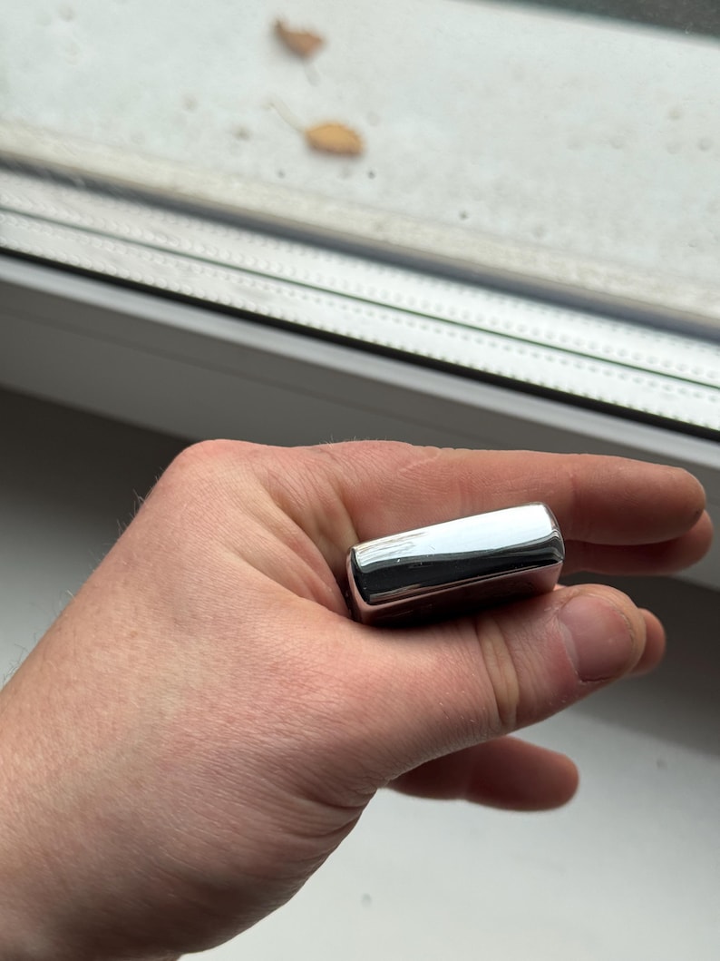May include: A shiny, rectangular, chrome-colored lighter held in a hand. The lighter has rounded edges and reflects the surrounding environment. The lighter is a classic design, suitable for lighting cigarettes or other flammable materials.