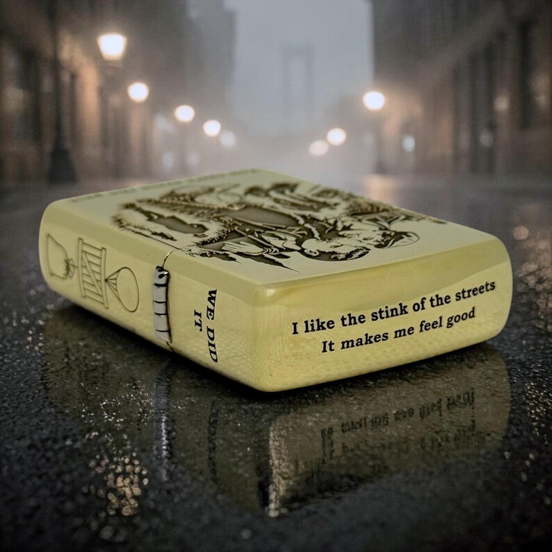 Pu&ograve; includere: Un accendino beige con un design inciso dettagliato e il testo "I like the stink of the streets. It makes me feel good." Il lato recita "WE DID IT". L'accendino &egrave; su una superficie bagnata.