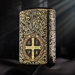 May include: A rectangular metal lighter with a brass finish featuring an intricate engraved scrollwork pattern. The front displays a Saint Benedict medal design with the text PAX, C S S M L, N D S M D, and the Latin phrase FIAT JUSTITIA ET PEREAT MUNDUS.