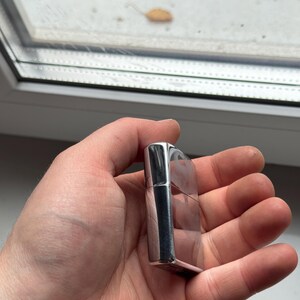 May include: A silver-colored, rectangular lighter with a hinged lid. The lighter has a polished, reflective surface and is held in a hand. The background shows a window with a blurred view.