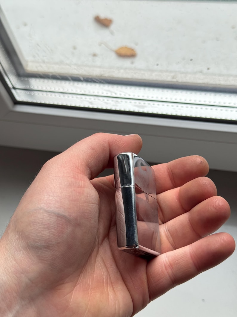 May include: A silver-colored, rectangular lighter with a hinged lid. The lighter has a polished, reflective surface and is held in a hand. The background shows a window with a blurred view.