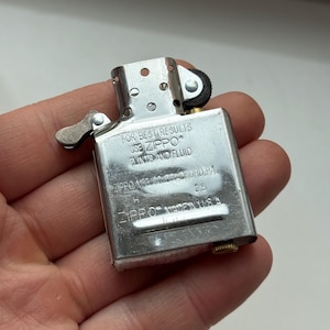 May include: A silver Zippo lighter with the text "FOR BEST RESULTS USE ZIPPO FLINTS AND FLUID" and "ZIPPO MADE IN U.S.A." The lighter is rectangular with a hinged lid and a small wheel for sparking. The lighter is held in a hand.