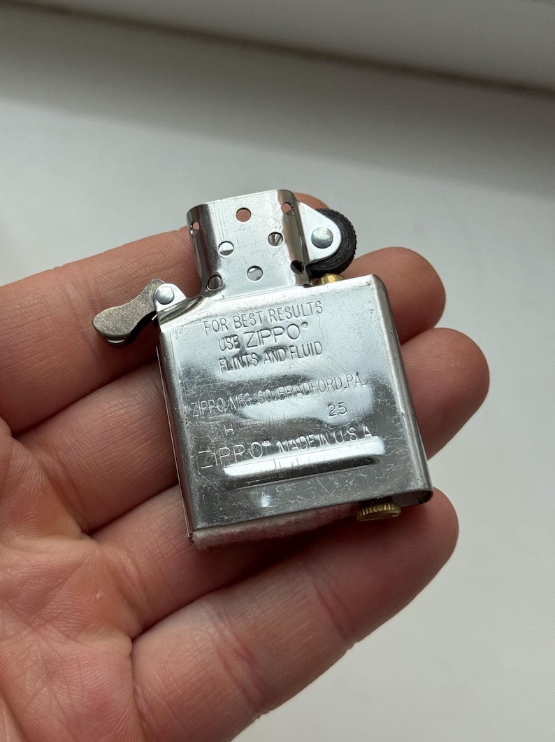 May include: A silver Zippo lighter with the text "FOR BEST RESULTS USE ZIPPO FLINTS AND FLUID" and "ZIPPO MADE IN U.S.A." The lighter is rectangular with a hinged lid and a small wheel for sparking. The lighter is held in a hand.