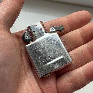 May include: A silver metal lighter with a hinged lid and a small lever. The lighter has text on it that reads "KEEP AWAY FROM CHILDREN, AFTER FILLING WIPE LIGHTER & HANDS BEFORE IGNITING, LIGHTER DOES, SELF EXTINGUISH, CLOSE LID TO PUT IT OUT."