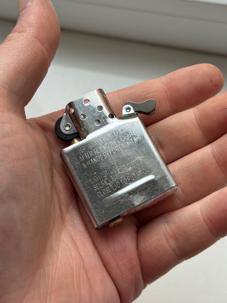 May include: A silver metal lighter with a hinged lid and a small lever. The lighter has text on it that reads "KEEP AWAY FROM CHILDREN, AFTER FILLING WIPE LIGHTER & HANDS BEFORE IGNITING, LIGHTER DOES, SELF EXTINGUISH, CLOSE LID TO PUT IT OUT."