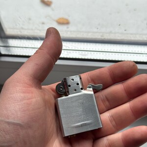 May include: A silver-colored metal lighter with a hinged lid and a small lever. The lighter has a rectangular body with a brushed metal finish and a series of small holes near the top. The lighter is held in a person's hand.