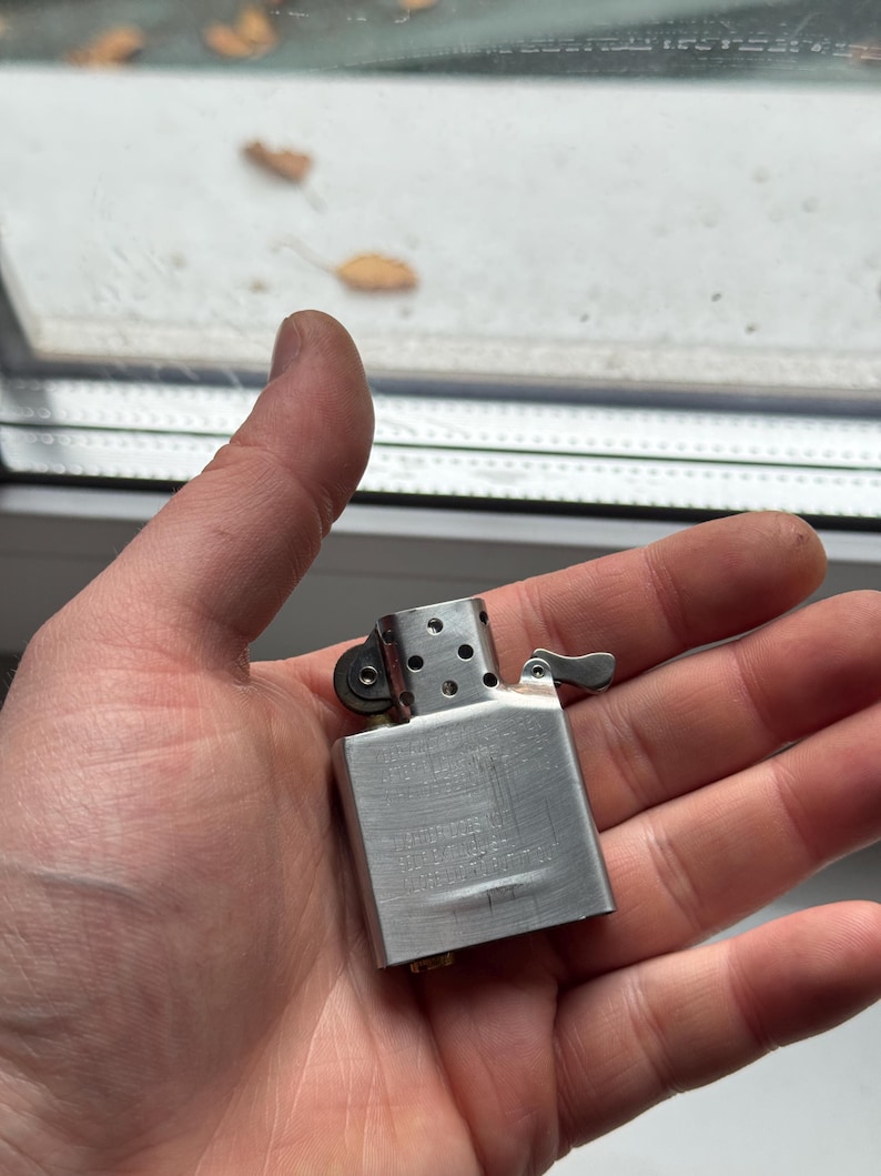 May include: A silver-colored metal lighter with a hinged lid and a small lever. The lighter has a rectangular body with a brushed metal finish and a series of small holes near the top. The lighter is held in a person's hand.