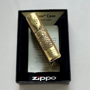 May include: A gold-colored Zippo lighter with an engraved skull design, presented in a black box with a blue interior. The lighter features intricate artwork and the Zippo logo.