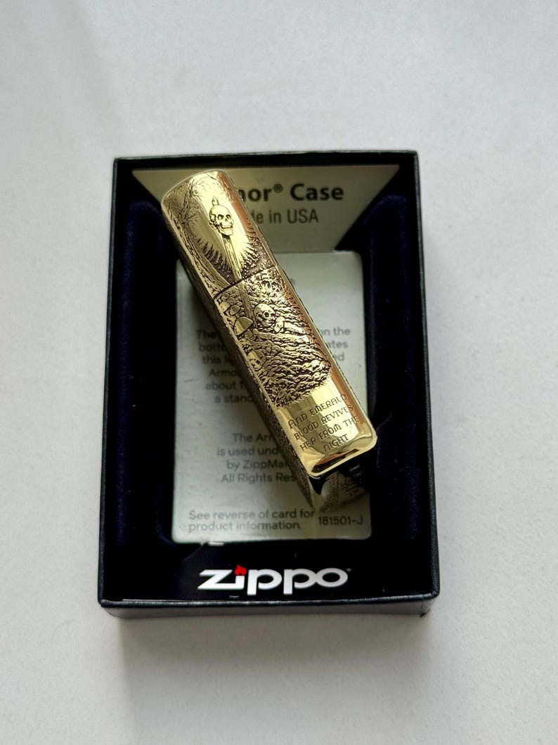 May include: A gold-colored Zippo lighter with an engraved skull design, presented in a black box with a blue interior. The lighter features intricate artwork and the Zippo logo.