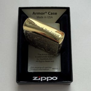 May include: A gold-colored Zippo Armor Case lighter with the text "Warden of the forest" engraved on the top. The lighter is in a black box with the Zippo logo and "Made in USA" printed on it.