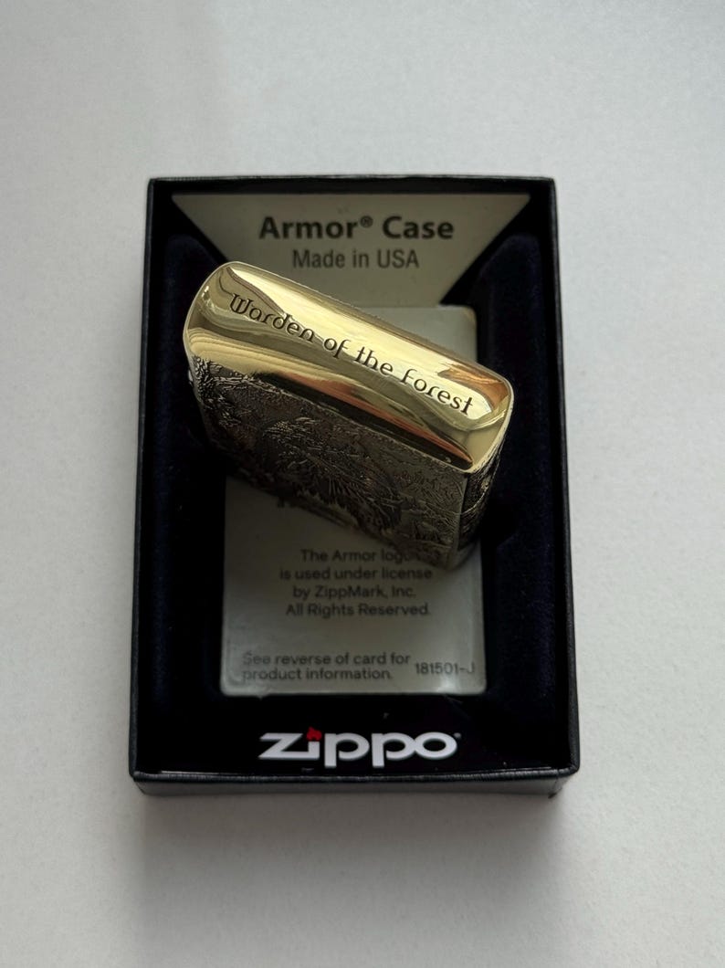 May include: A gold-colored Zippo Armor Case lighter with the text "Warden of the forest" engraved on the top. The lighter is in a black box with the Zippo logo and "Made in USA" printed on it.