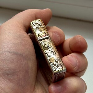 May include: A silver and gold-toned lighter with an engraved wave and floral pattern. The lighter is held in a hand and has a hinged lid. The design features intricate details and a vintage aesthetic.