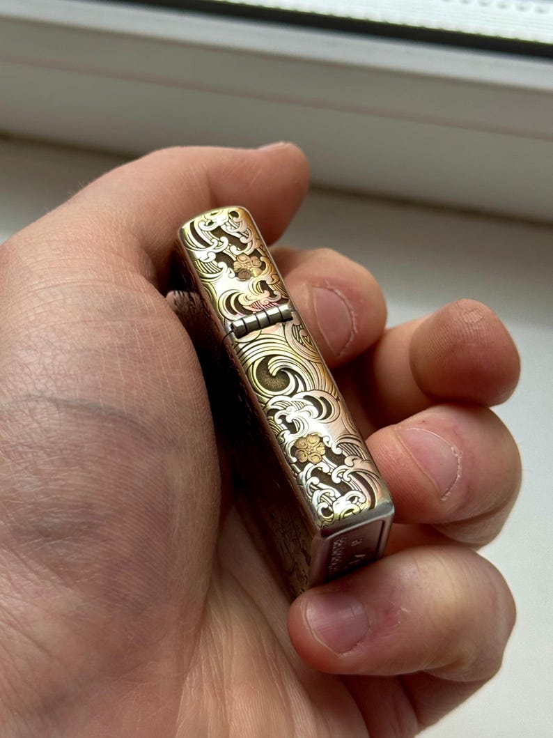 May include: A silver and gold-toned lighter with an engraved wave and floral pattern. The lighter is held in a hand and has a hinged lid. The design features intricate details and a vintage aesthetic.