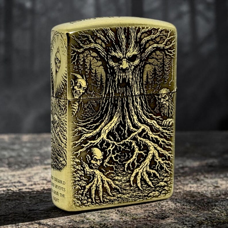 May include: A brass-colored lighter with an intricate engraved design. The design features a gnarled tree with a face, surrounded by forest imagery and grotesque figures. The lighter has text on the side that reads "Emerald...revives...from the...night."