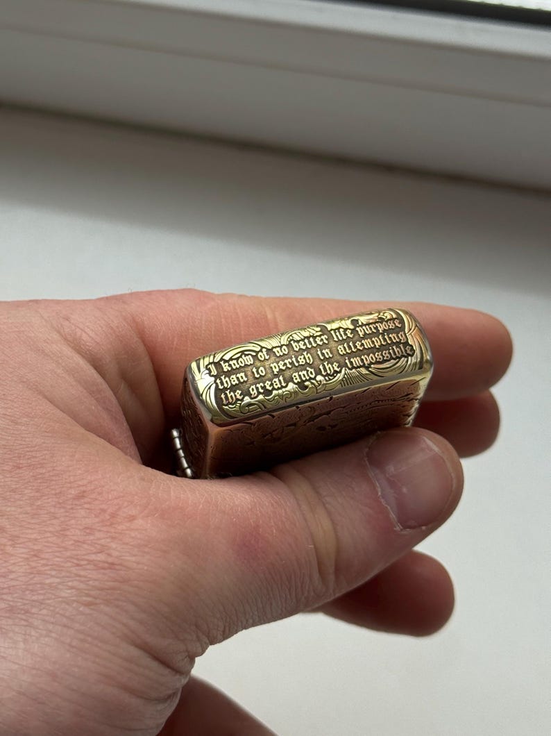 May include: A gold-colored metal lighter with engraved text: "I know of no better life purpose than to perish in attempting the great and the impossible." The lighter has a rectangular shape and a chain.