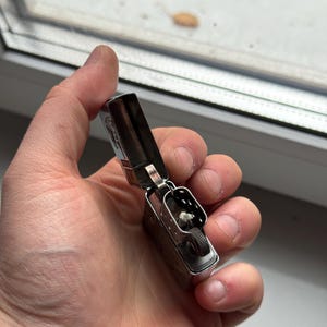 May include: A silver metal lighter with the top open, revealing the wick and flint wheel. The lighter is held in a hand, and the background shows a window and some leaves.