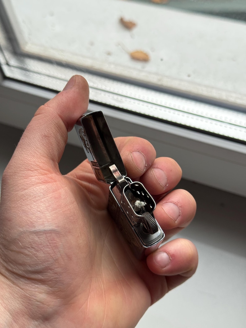 May include: A silver metal lighter with the top open, revealing the wick and flint wheel. The lighter is held in a hand, and the background shows a window and some leaves.