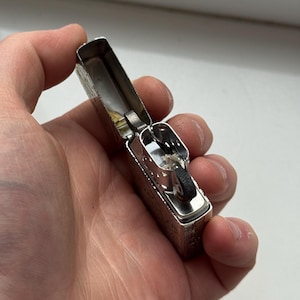 May include: Silver metal lighter with an open lid, revealing the internal components. The lighter has an engraved design on the exterior. The lighter is held in a person's hand.