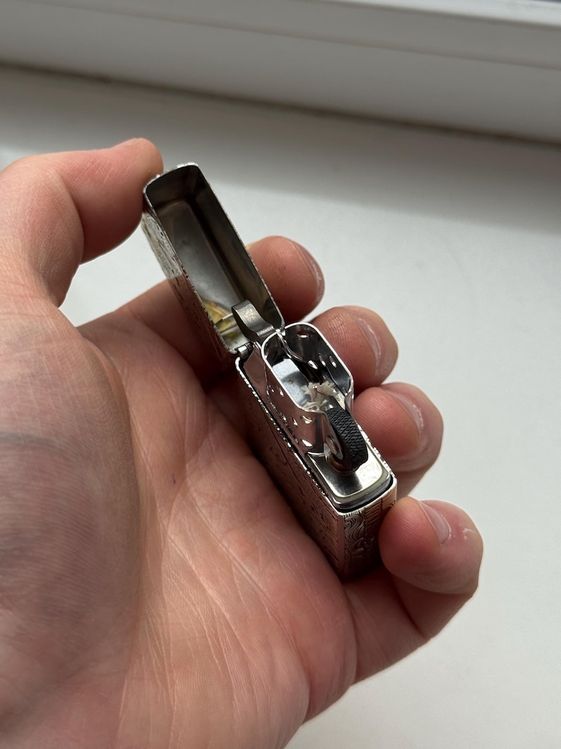 May include: Silver metal lighter with an open lid, revealing the internal components. The lighter has an engraved design on the exterior. The lighter is held in a person's hand.