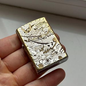 May include: A gold and silver metal lighter with an intricate dragon design. The lighter has a rectangular shape with rounded edges. The dragon design covers the entire surface of the lighter, with detailed scales and features.