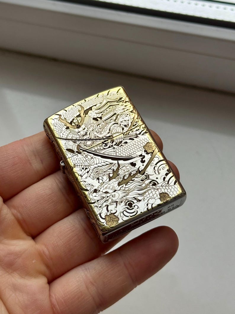 May include: A gold and silver metal lighter with an intricate dragon design. The lighter has a rectangular shape with rounded edges. The dragon design covers the entire surface of the lighter, with detailed scales and features.