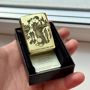 Pu&ograve; includere: Un accendino color oro con un design inciso raffigurante una coppia, paesaggi urbani e un orologio da tasca. Il testo "You can't erase the past" &egrave; visibile. L'accendino &egrave; in una scatola nera.