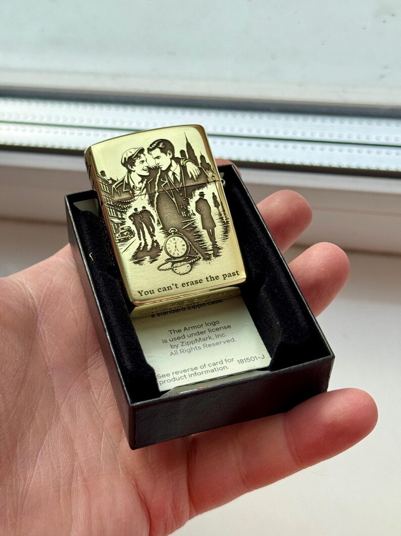 Pu&ograve; includere: Un accendino color oro con un design inciso raffigurante una coppia, paesaggi urbani e un orologio da tasca. Il testo "You can't erase the past" &egrave; visibile. L'accendino &egrave; in una scatola nera.