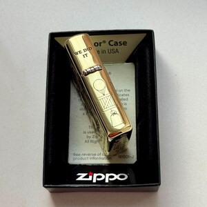 Pu&ograve; includere: Un accendino Zippo color oro in una scatola nera. L'accendino presenta la scritta "WE DID IT" incisa, insieme ad altri disegni. Il logo Zippo &egrave; visibile sulla scatola.