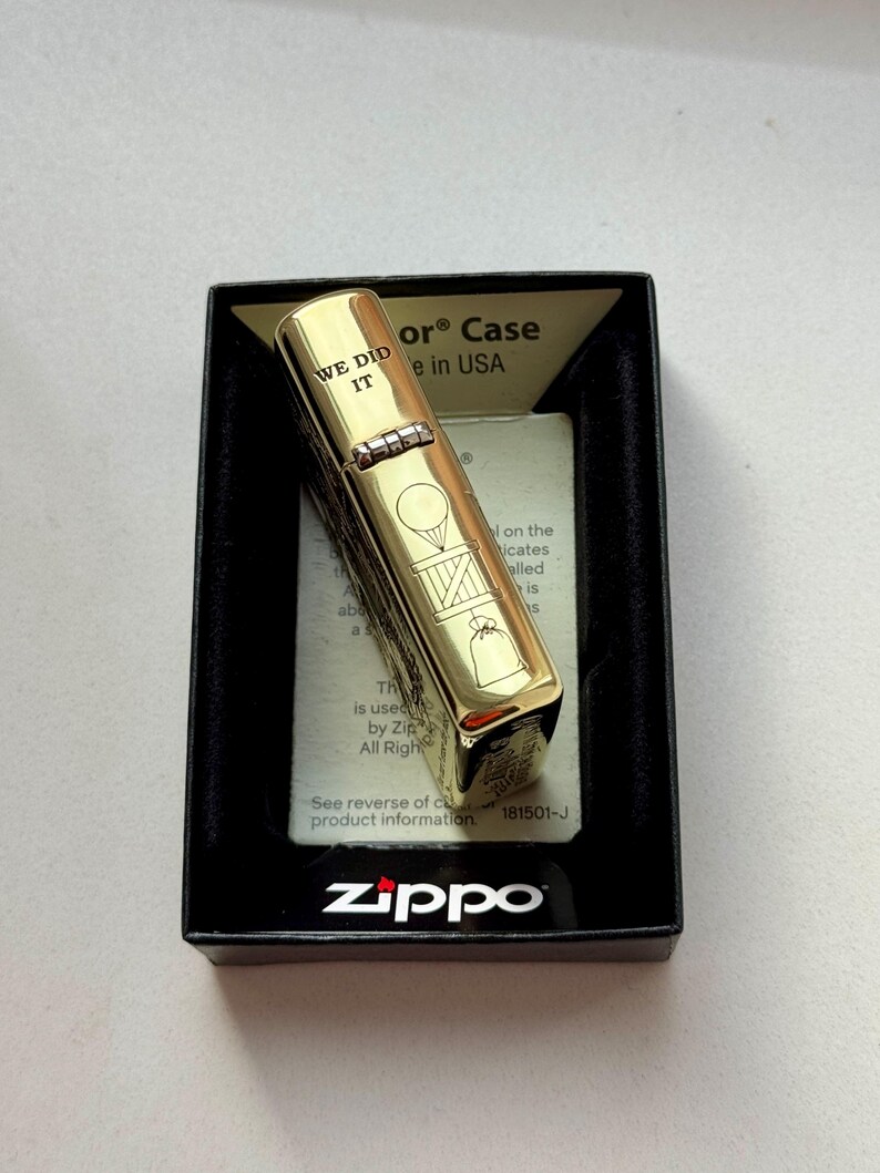 Pu&ograve; includere: Un accendino Zippo color oro in una scatola nera. L'accendino presenta la scritta "WE DID IT" incisa, insieme ad altri disegni. Il logo Zippo &egrave; visibile sulla scatola.