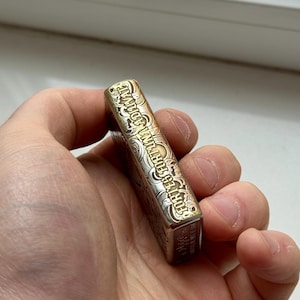 May include: A rectangular, metallic lighter with ornate engravings. The lighter has a gold and silver finish, with the text "FORTES FORTUNA ADIUVAT" etched along the side. The lighter is held in a hand.